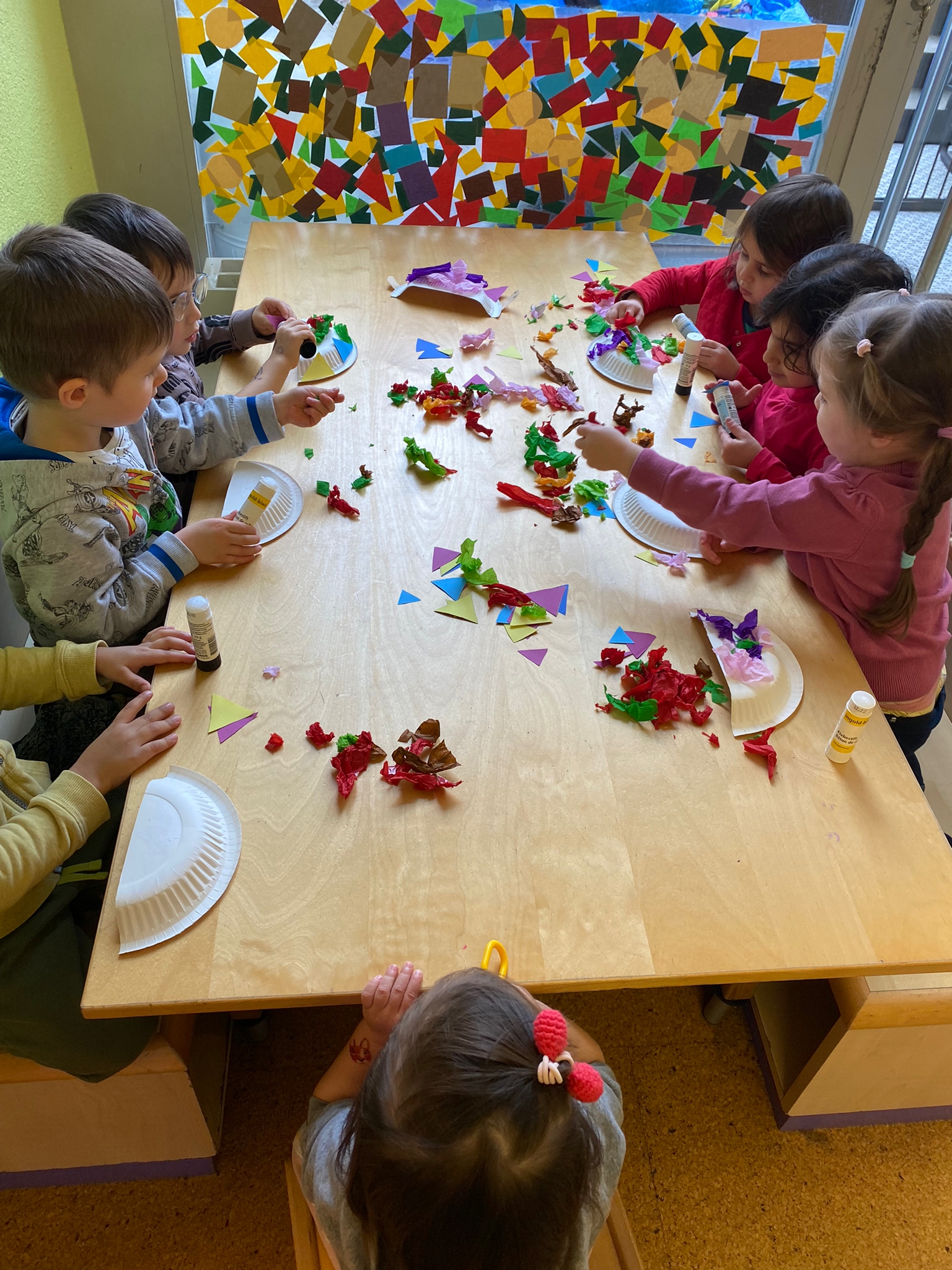 Children creating colourful paper collages at a table covered in torn tissue paper and shapes