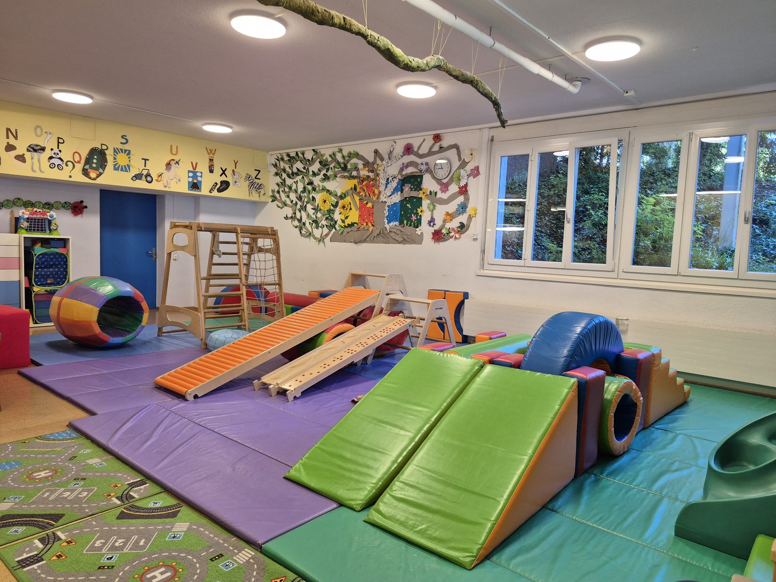 Bright movement and soft-play room with climbing frames, ramps, gymnastic mats and an alphabet display along the wall